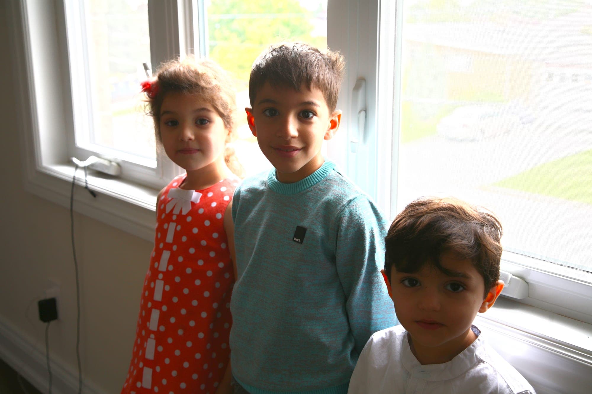 Three young children posed together by a bright window