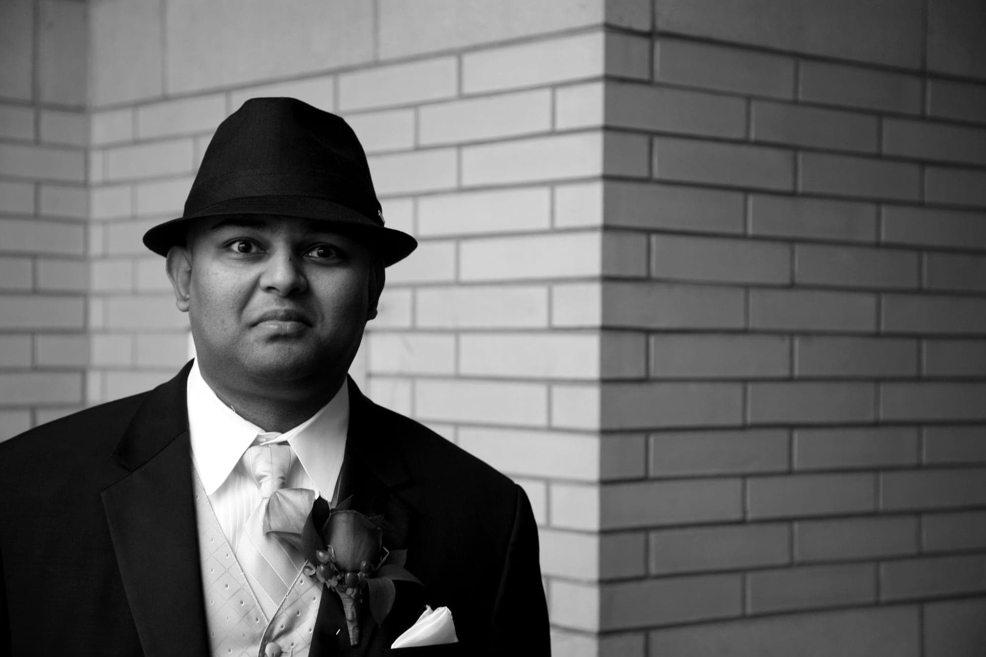 Black and white editorial portrait of a groom in a fedora against brick