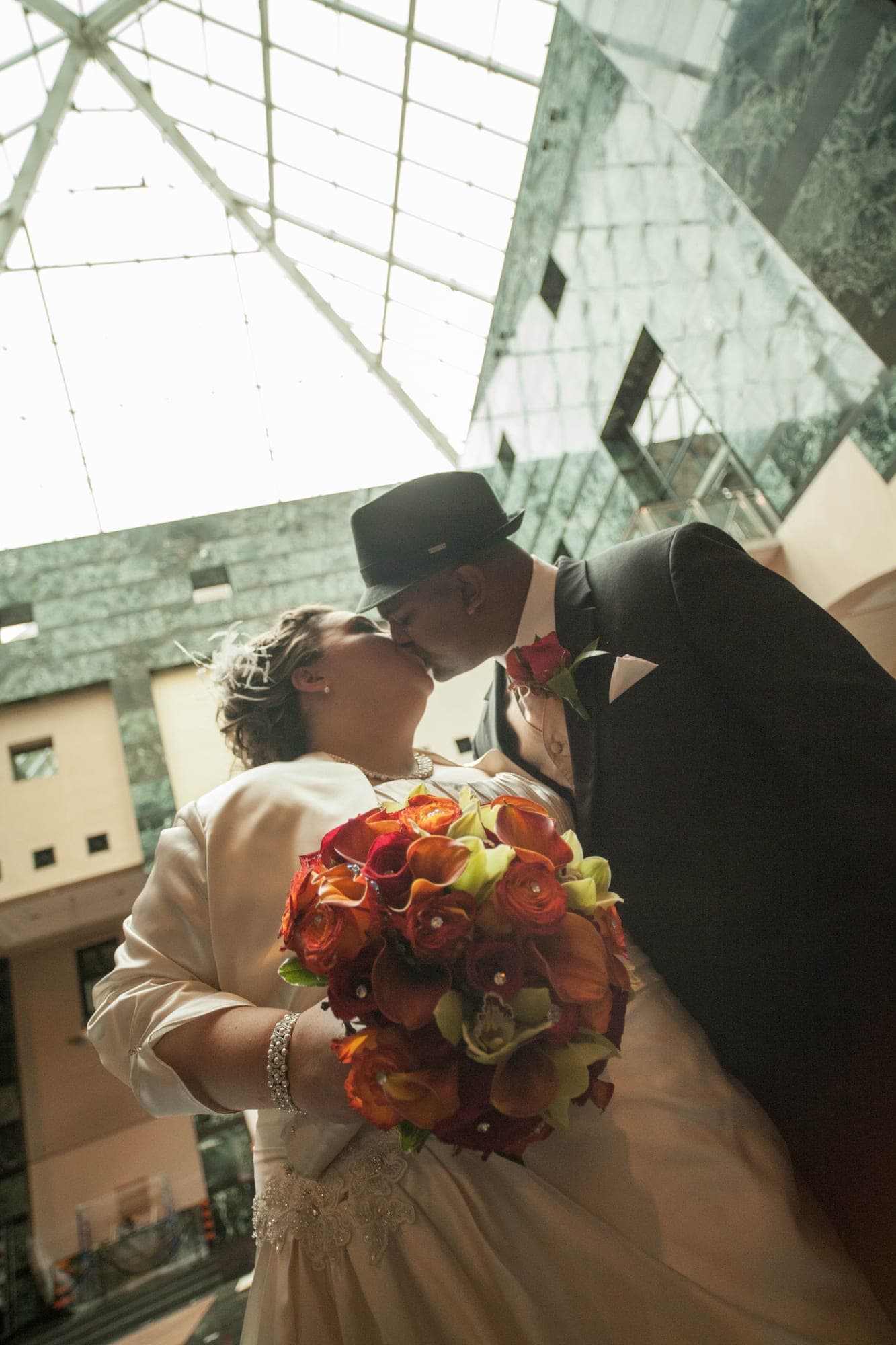 Bride and groom kissing beneath a glass atrium, bouquet raised